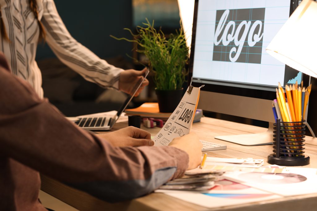 Detailaufnahme eines Schreibtisches. Auf diesem steht ein PC, der das Wort "Logo" zeigt. An dem Tisch sitzt und steht ein Mensch. Die beiden Personen arbeiten an einem gemeinsamen Projekt und sind nur bis zum Oberkörper erkennbar. Die vordere Personen hält ein Blatt leicht hoch vom Tisch und während die andere ein weiteres kleines Laptop dahinter öffnet.