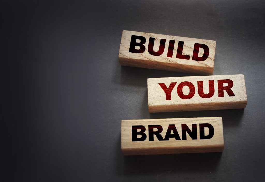 Drei Klötze liegen auf einem grauen Hintergrund. Auf jedem Klotz steht jeweils ein Wort. AUf dem ersten steht "Build", auf dem Zweiten "your" und auf dem Dritten "Brand". Alle Holzklätze sind versetzt untereinander gelegt und finde sich allesamt in der rechten Bildhälfte.