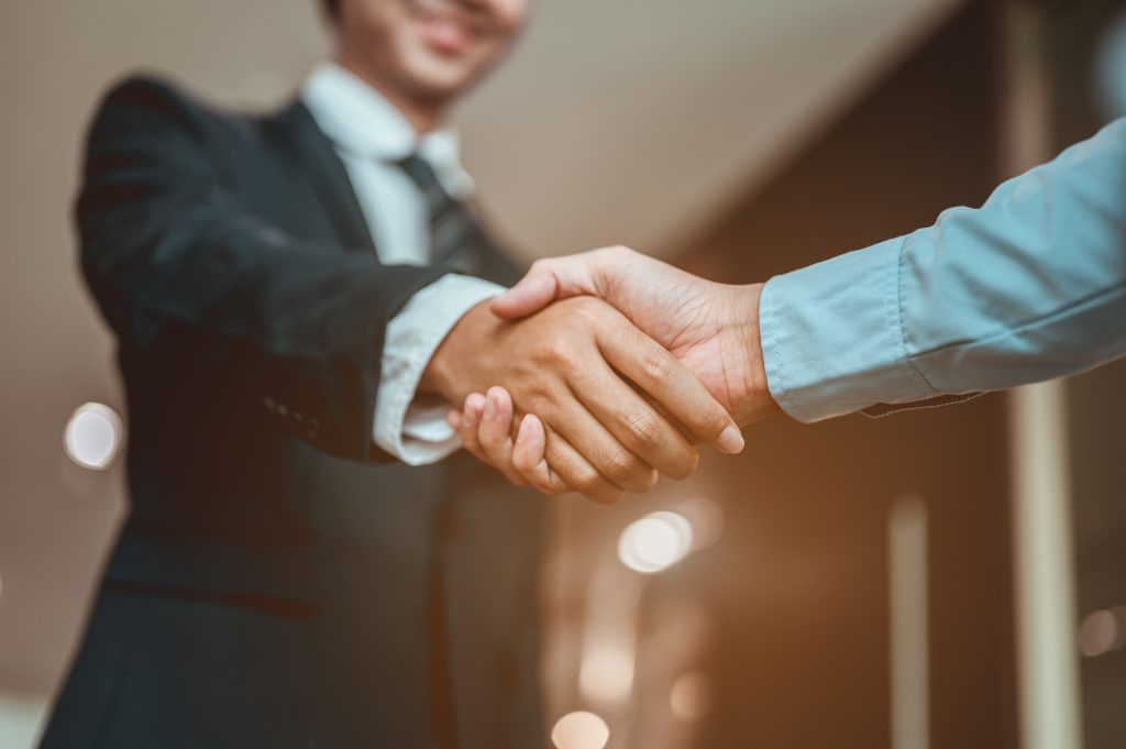 Zwei Personen schütteln sich die Hand. Von der vorderen Personen ist nur die Hand zu sehen, von der hinteren Personen erkennt man auch einen Teil des Oberkörpers und Halses. Man schaut aus einer leichten Froschperspektive auf die Situation.