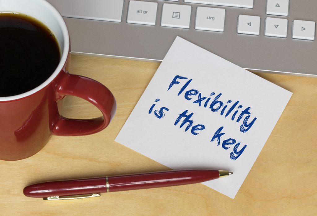 Detailaufnahme von einer Tasse auf der linken Seite, einem Laptop dahinter am oberen Bildrand, einem Zettel in der Mitte mit der Aufschrift "Flexibility is the key" und einem Stift, der am unteren Bildrand halb auf dem viereckigen Papierzettel liegt.