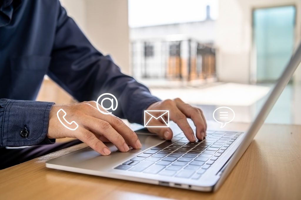 Mensch sitzt an einem Holztisch und arbeitet am Laptop. Man sieht nur eine Detailaufnahme der Hände und des Laoptops während der Arbeit. Über dem Laptop leuchten viele kleine Icons auf, z.B. ein @-Icon, ein E-Mail Icon und ein Telefon Icon.